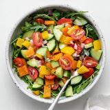 A top-down view of a white speckled ceramic bowl filled with a colorful mixed green salad, including arugula, cucumbers, tomatoes, peppers, and onions, with a metal spoon resting inside.