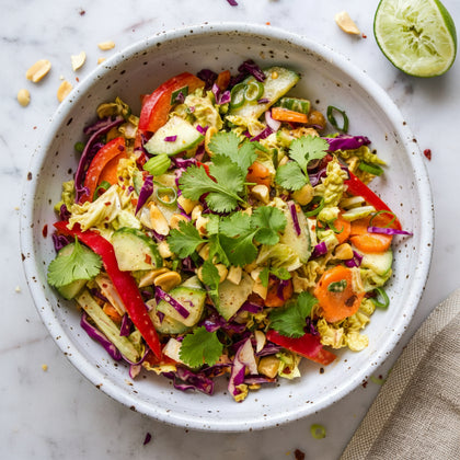 A close-up overhead shot of a colorful salad in a speckled white bowl, featuring avocado, red cabbage, cucumber, carrots, red bell pepper, and peanuts, with a lime half visible in the top right on a light marble surface.