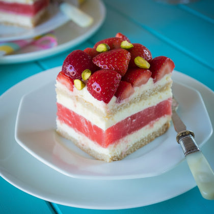 A slice of strawberry and cream cake on a white plate with a fork, set on a turquoise table with another slice blurred in the background.